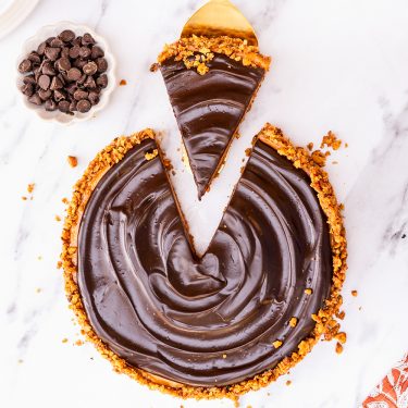 Chocolate tart with a nutty crust, one slice removed, on a marble surface. A small bowl of chocolate chips is next to the tart.