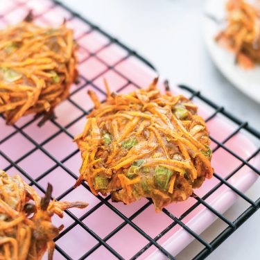 Crispy vegetable fritters made with shredded carrots and green onions are cooling on a black wire rack over a pink tray.
