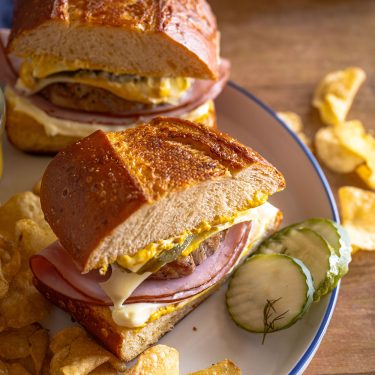 Sandwich with ham, cheese, and mustard on crusty bread, served with pickles and potato chips on a plate.