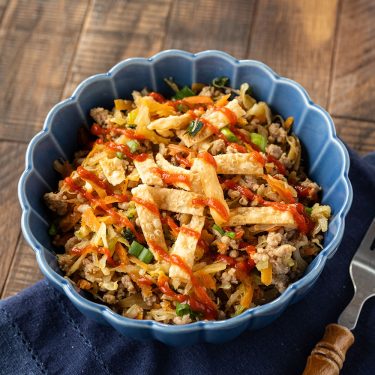 A blue bowl filled with an egg roll in a bowl, topped with crispy strips, green onions, and a drizzle of sriracha sauce, on a wooden table.