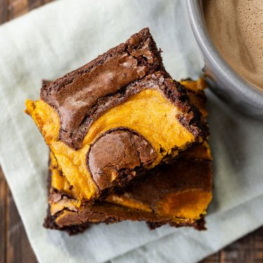 Two pumpkin swirl brownies stacked on a napkin beside a cup of coffee.