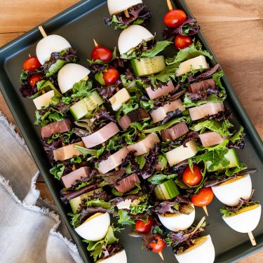 Skewers with boiled eggs, lettuce, cherry tomatoes, cucumber, cheese, and ham arranged neatly on a black tray.
