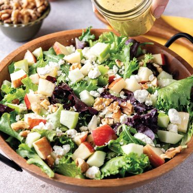 Mixed greens salad with diced apples, walnuts, and crumbled cheese in a wooden bowl, with a hand pouring dressing from a jar.