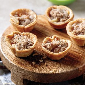 Five mini apple crumble tarts with golden crusts and crumbly toppings displayed on a round wooden board.