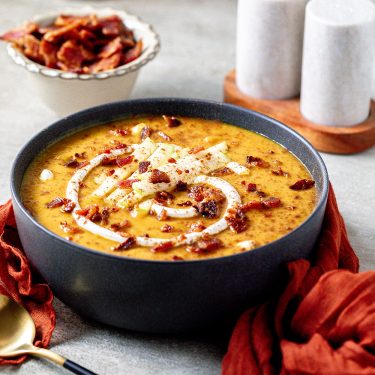 A bowl of creamy soup topped with bacon bits and thinly sliced vegetables, with a swirl of cream, a gold spoon, and a side bowl of bacon in the background.