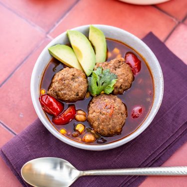 Bowl of soup with meatballs, sliced avocado, red bell peppers, corn, chickpeas, and cilantro garnish, placed on a purple napkin with a spoon nearby.