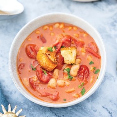 A bowl of tomato and bean soup with chunks of bread and fresh herbs, served in a white bowl on a marble surface.