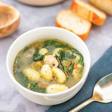 A bowl of chicken gnocchi soup with spinach, served with slices of bread on the side and a spoon nearby.