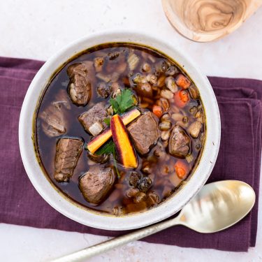 A bowl of beef and vegetable soup with chunks of beef, carrots, and barley, garnished with parsley, on a purple napkin with a spoon beside it.