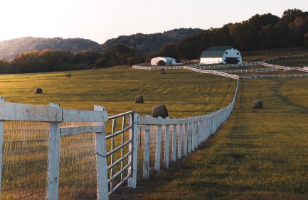 A rural farm landscape with white fences, green fields, hay bales, two barns, and trees in the background during sunset.