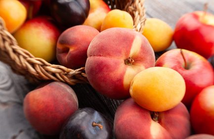 A close-up of assorted fresh stone fruits, including peaches, apricots, plums, and apples, spilling out of a woven basket onto a wooden surface.